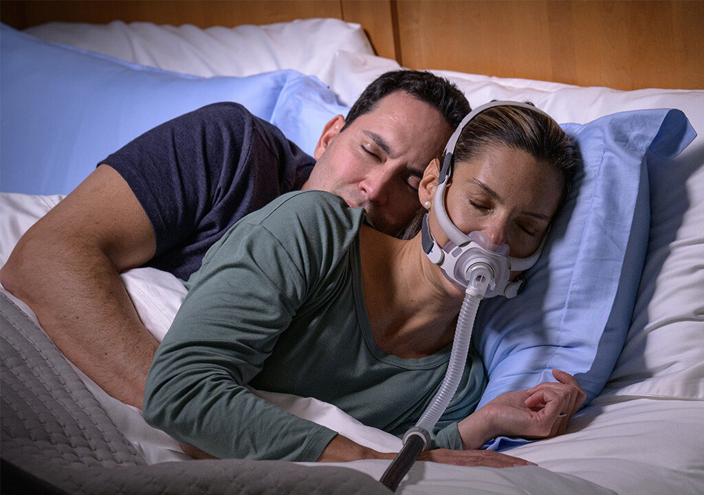 Un hombre y una mujer acostados y durmiendo en una cama, la mujer usa una mascarilla AirFit F40