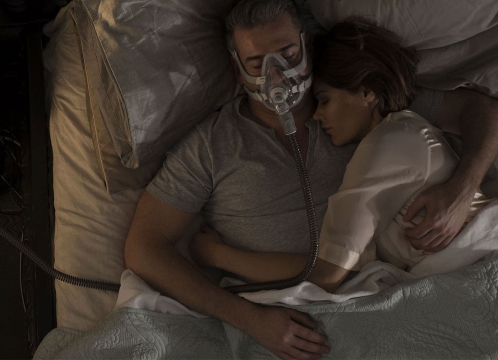 Dos personas duermen abrazadas, el hombre usa una mascarilla para tratamiento de apnea