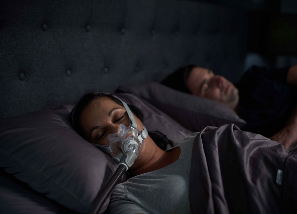 Dos personas duermen en una cama, la mujer en primer plano usa una mascarilla para tratamiento de apnea
