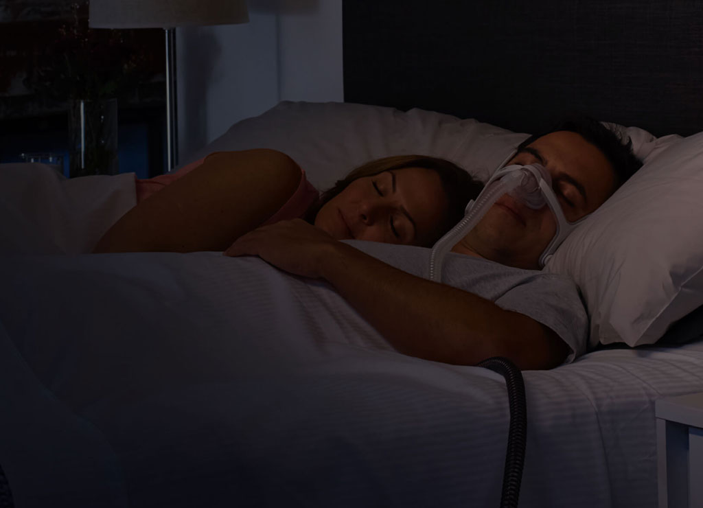Un hombre acostado en una cama con una persona duerme mientras usa la mascarilla AirFit N20