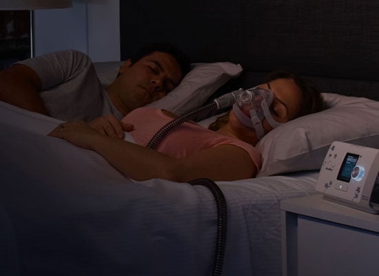 Una pareja duerme mientras la mujer usa la mascarilla AirTouch F20 de la marca ResMed