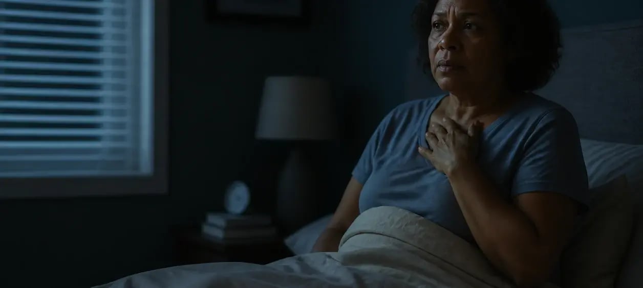 Mulher sentada na cama durante a noite, com expressão de preocupação e a mão sobre o peito.